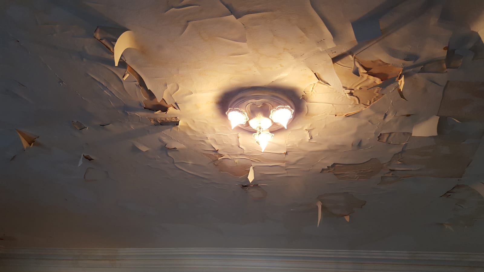 Severely water-damaged ceiling with paint and plaster peeling in sheets around a light fixture